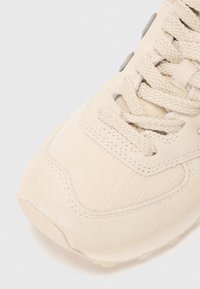 Close-up of beige sneaker toe with textured fabric, suede details, and thick woven laces on a light background.