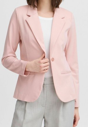 Kvinde iført en lyserød blazer, hvid top og lysegrå bukser, holder blazeren lukket med den ene hånd.
