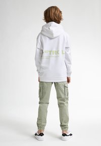 Felpa bianca con logo nero in verde chiaro, abbinata a pantaloni cargo verdi chiari. Presenta polsini elastici e sneakers bianche con dettagli neri.