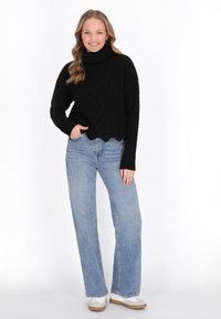 Schwarzer Strickpullover mit Rollkragen und Zopfmuster, kombiniert mit hellblauen, hoch-taillierten gerade geschnittenen Jeans und weißen Schuhen.