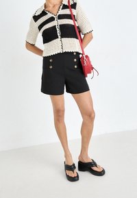Czarno-biała bluzka dzianinowa w paski z falbankami na brzegach, zestawiona z czarnymi szortami z wysokim stanem z złotymi guzikami i czerwoną torbą.