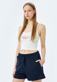 Weißes geripptes Crop-Top mit rotem "MANHATTAN"-Druck, kombiniert mit dunkelblauen Shorts mit elastischem Bund. Einfaches Design, keine Muster oder zusätzlichen Details.
