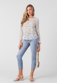 White lace top with floral cutouts, long sleeves, paired with light blue ankle-length jeans. Model holds a small quilted handbag and wears beige heels.
