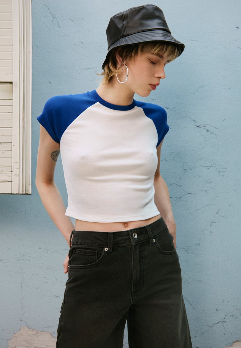 Maglietta bianca corta con maniche azzurre a raglan, abbinata a jeans neri a vita alta. Lo sfondo è una parete blu conciata.