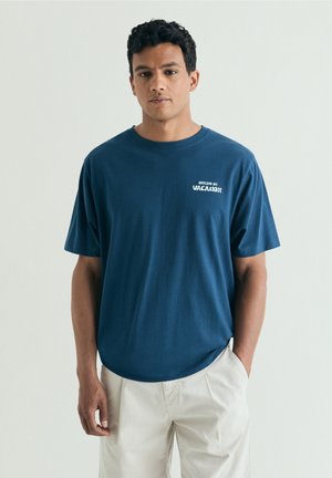 Bărbat purtând un tricou albastru cu textul „Offline în vacanță”, combinat cu pantaloni bej deschis, stând în fața unui fundal simplu și deschis la culoare.