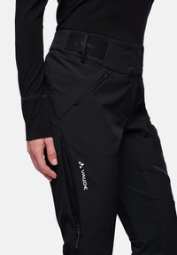 Zwarte softshell broek met verstelbare tailleband, ritszakken aan de zijkant en textuurelementen. Bevat een opvallend wit logo.