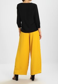 Top nero a tre quarti abbinato a pantaloni gialli a gamba larga, con una texture liscia e una silhouette pulita e moderna.