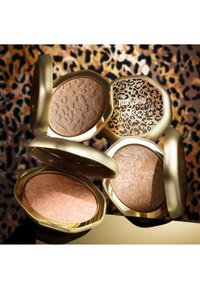 Quatre poudriers compacts ouverts de ton doré avec des surfaces texturées et des couvercles imprimés léopard sur un fond assorti imprimé léopard.
