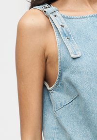 Gros plan d'une personne portant une salopette en denim bleu clair avec une bretelle ajustable et une boucle argentée sur une épaule nue, sur un fond blanc.