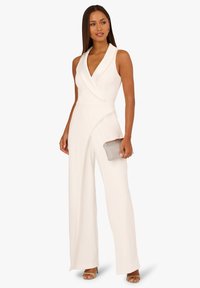 Bílý overal s hlubokým výstřihem do V, zavinovacím střihem a ozdobným lemováním. Široké nohavice s jemným leskem, doplněný malou stříbrnou kabelkou.