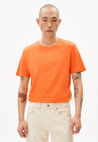 T-shirt en coton orange à manches courtes avec un col rond et une texture douce, porté avec un jean clair. Présente des tatouages visibles.