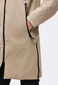Veste longue beige avec des fermetures éclair noires, une main dans la poche latérale zippée, et un pantalon noir visible en bas.