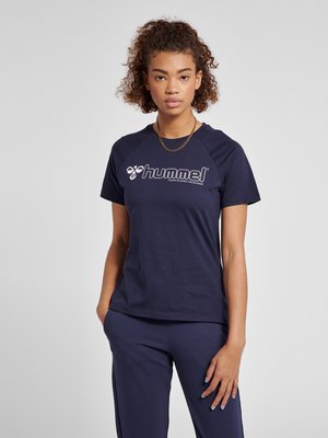 Jonge vrouw met krullend haar die een marineblauw hummel t-shirt en bijpassende broek draagt, staand met één hand in de zak tegen een effen achtergrond.