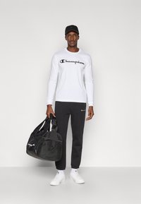 Λευκό πουκάμισο με μακριά μανίκια και μαύρο λογότυπο "Champion", μαύρο παντελόνι φόρμας, μαύρη τσάντα duffel και λευκά αθλητικά παπούτσια· μινιμαλιστικός αθλητικός σχεδιασμός.