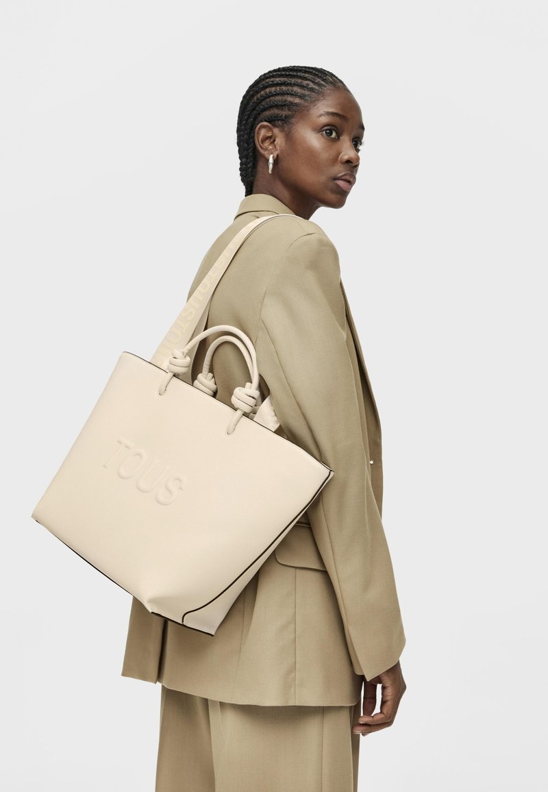 Beżowa skórzana torba na ramię z wytłoczonym logo "TOUS", dwoma górnymi uchwytami i wzorzystym paskiem, z kontrastującymi czarnymi krawędziami.