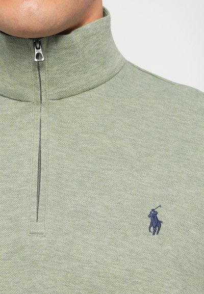 Ανοιχτό πράσινο πουλόβερ με υψηλό γιακά, ημικλείσιμο φερμουάρ και μπλε κεντητό λογότυπο. Μαλακό, υφασμάτινο υλικό με διακριτό σχέδιο.