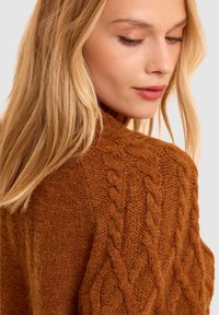 Maglione a maglia trecciata in arancione bruciato, con colletto alto e motivo testurizzato. Materiale morbido con dettagli di cucitura ben visibili.