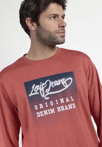 Camisa de manga larga, de color rojizo, con un diseño gráfico que presenta el logo "Lois Jeans" y el texto "Original Denim Brand" en blanco sobre un fondo azul oscuro.