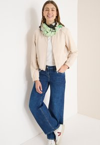 Giacca bomber beige con polsini elasticizzati, t-shirt bianca, jeans larghi blu e sciarpa verde con motivi. Sneakers bianche con dettagli rossi.