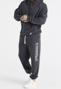 Persoon in een donkergrijze hoodie en bijpassende joggers met witte patroon "SikSilk" tekst op het been, en witte sneakers met een gouden logo.