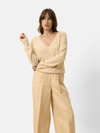 Beige V-Ausschnitt-Pullover aus weichem Material, kombiniert mit locker sitzenden beigen Hosen. Das Outfit zeichnet sich durch ein entspanntes Design und eine glatte Textur aus.