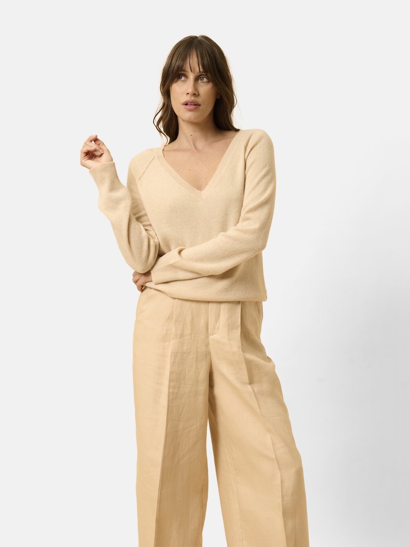 Beige V-Ausschnitt-Pullover aus weichem Material, kombiniert mit locker sitzenden beigen Hosen. Das Outfit zeichnet sich durch ein entspanntes Design und eine glatte Textur aus.