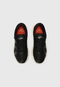 Svart sneakers med strukturert materiale, kontrasterende hvit såle og rød merkevaredetalj inni. Snørebånd, polstret krage og logo detaljer til stede.
