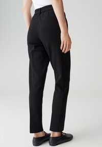 Personne portant un pantalon noir slim, des chaussures noires à enfiler et un haut blanc, vue de dos sur un fond clair uni.