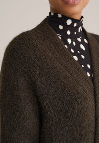 Smeđi pleteni kardigan s teksturiranim, dlačicama sličnim završetkom i dubokim V izrezom. Nosite ga preko crnog rolka majice s uzorkom bijelih točkica.