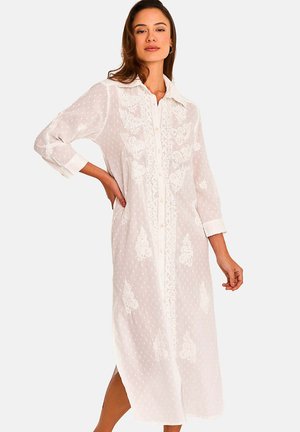Femme portant une longue robe blanche boutonnée en tissu transparent avec une broderie florale complexe, posant avec une main sur la hanche et un fond neutre.