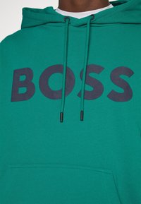 Sweat à capuche vert avec des cordons de serrage noirs et un grand texte noir "BOSS" sur la poitrine, porté par une personne à la peau foncée.