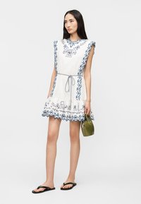 Γυναίκα με λευκό αμάνικο φόρεμα με μπλε κεντήματα και κυματιστές άκρες, κρατώντας μια τσάντα ελιάς, φορώντας μαύρα σανδάλια.