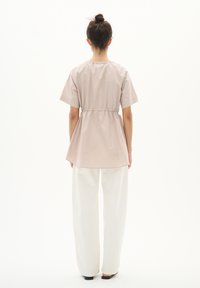 Lys beige kortærmet top med elastisk talje, let udvidet silhuet og glat, skinnende tekstur. Kombinationen er med hvide bredbenede bukser.