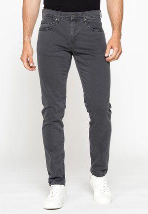 Uomo che indossa jeans aderenti grigio scuro e sneakers bianche in piedi davanti a uno sfondo chiaro e uniforme.