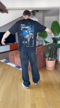 Fekete póló kék virágos grafikával és felirattal, laza fekete farmerrel párosítva. A háttérben egy növény és egy barna kanapé látható. Fapadló.