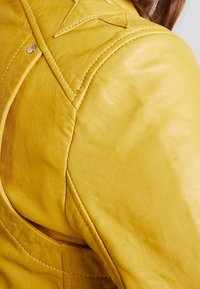 Giacca di pelle gialla con texture liscia, cuciture in evidenza, accento a forma di stella sulla spalla e dettagli con bottoni a pressione argentati.