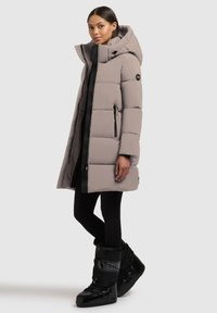 Abrigo acolchado en gris claro, con capucha, bolsillos con cremallera y detalles en negro. Lleva puesto sobre un outfit negro y combinado con botas negras.