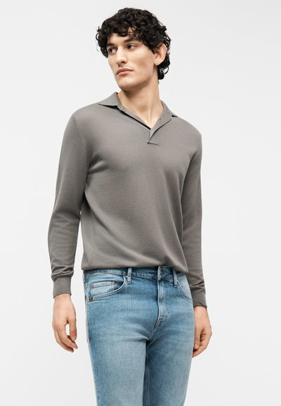 Νεαρός άνδρας με κυματιστά μαλλιά, ντυμένος με γκρι μακρυμάνικο πόλο και ανοιχτόχρωμα τζιν, που κοιτάζει στο πλάι με φόντο απλό.