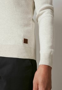 Light beige sweater with ribbed cuff and hem. Features a small brown leather patch on the lower side. Soft texture, slim fit design.