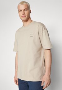 Beige bomull T-skjorte med korte ermer, med to svarte "SAMSOE"-logoer på brystet. Klassisk passform med rund hals og et enkelt design.