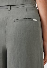 Světle šedé kalhoty s hladkým, texturovaným povrchem. Obsahují malou zadní kapsu a štítek označený "ALLSAINTS." Střih přizpůsobený postavě s čistou siluetou.