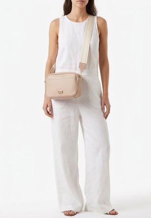 Femme portant une combinaison blanche sans manches à jambes larges, portant un sac beige bandoulière avec une large sangle sur l'épaule gauche.