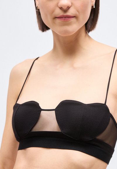 Γυναίκα που φοράει μαύρο σουτιέν με λεπτές τιράντες και διάτρητο πάνελ μεταξύ των υφασμάτινων κυπέλλων σε σχήμα καρδιάς, σε ανοιχτό φόντο.