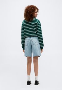 Γυναίκα με σγουρά μαλλιά που φοράει μακρυμάνικη μπλούζα με πράσινες και λευκές ρίγες, ανοιχτόχρωμα τζιν σορτς, λευκές κάλτσες και μαύρα παπούτσια, με την πλάτη γυρισμένη.