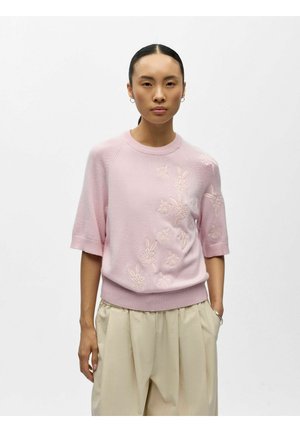 Jeune femme aux cheveux lisses portant un pull à manches courtes rose clair avec une broderie florale et un pantalon beige à jambes larges.