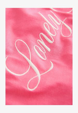 Roosa kangas, millel on sile tekstuur ja valge tikandiga kiri "Lonely" voolavas kaligraafilises stiilis.