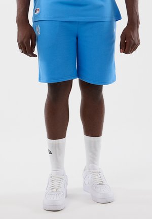 Uomo con pantaloncini blu vivace dei New York Yankees, calzini bianchi alla caviglia e sneaker bianche Nike in piedi contro uno sfondo semplice.