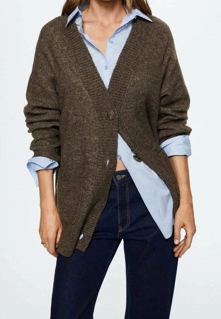 Femme portant un cardigan marron boutonné par-dessus une chemise bleu clair aux manches retroussées et un jean bleu foncé, sur un fond blanc.