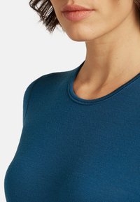 Woman with short dark hair wearing a teal crewneck shirt, showing lower face, neck, and upper chest on a white background.