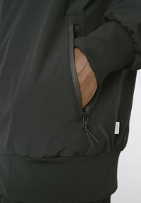 Schwarze Außenjacke mit glatter, wasserdichter Textur; verfügt über eine Reißverschlusstasche, gerippte Bündchen und ein Logoetikett am Saum.